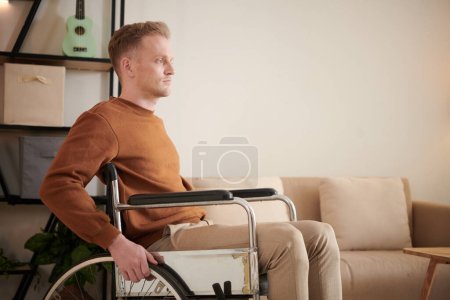 Téléchargez les photos : Sérieux jeune homme handicapé se déplaçant dans la maison en fauteuil roulant - en image libre de droit