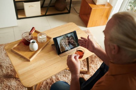 Téléchargez les photos : Homme âgé avec tablette numérique boire du thé et de la vidéo appelant sa fille et petits-enfants - en image libre de droit