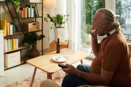 Téléchargez les photos : Homme âgé manger un sandwich et regarder l'émission populaire à la télévision à la maison - en image libre de droit