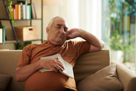 Téléchargez les photos : Homme âgé s'est endormi sur le canapé dans le salon lors de la lecture de roman - en image libre de droit