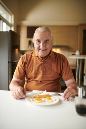 Téléchargez les photos : Portrait de l'homme âgé heureux ayant deux oeufs frits et sandwich pour le petit déjeuner à la maison - en image libre de droit