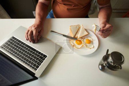 Foto de Hombre mayor comiendo huevos para el desayuno y leyendo noticias en la pantalla del ordenador portátil, vista desde arriba - Imagen libre de derechos