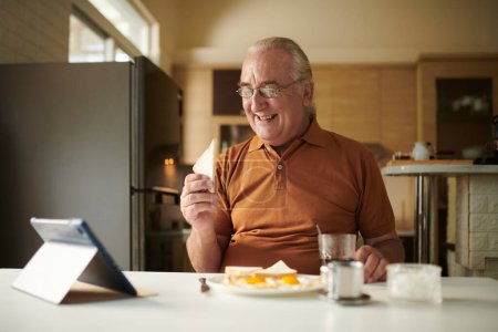 Foto de Alegre hombre mayor desayunando y viendo programa de comedia en la tableta de la computadora en casa - Imagen libre de derechos