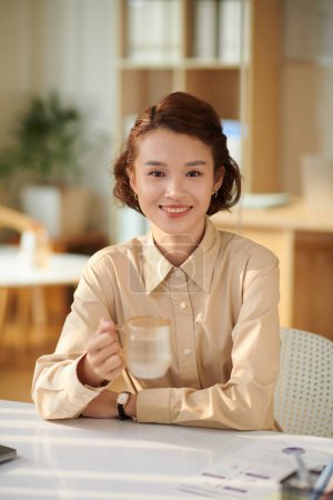 Photo for Portrait of smiling young businesswoman drinking cup of tea at office desk during break - Royalty Free Image