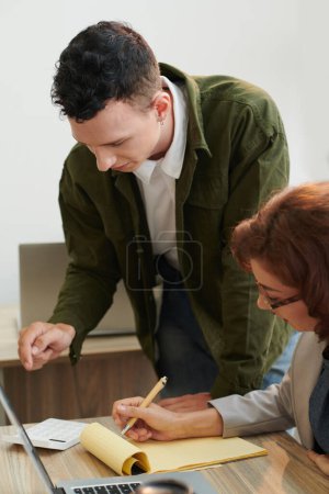 Téléchargez les photos : Jeune homme d'affaires faisant des calculs lorsque son collègue écrit dans le planificateur - en image libre de droit