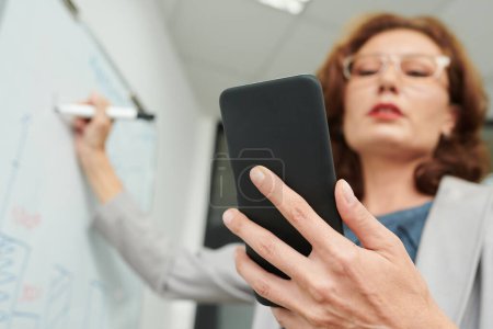 Téléchargez les photos : Femme entrepreneur mature vérifiant le document sur l'écran du smartphone lors de l'écriture sur tableau blanc - en image libre de droit