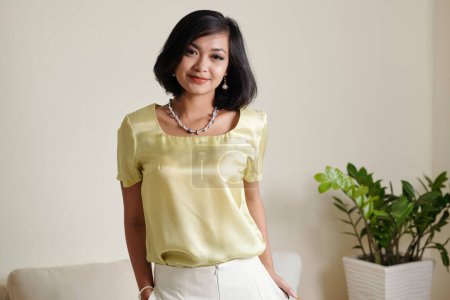 Téléchargez les photos : Portrait de positive jeune femme asiatique en satin blouse debout dans la chambre lumineuse - en image libre de droit