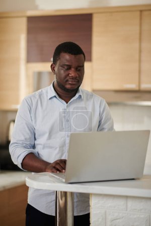 Téléchargez les photos : Homme noir sérieux debout au comptoir de la cuisine et de vérifier les chats de travail sur ordinateur portable - en image libre de droit