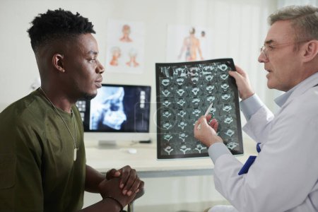 Photo for Serious doctor pointing at MRI image of spine when talking to military man suffering from back pain - Royalty Free Image