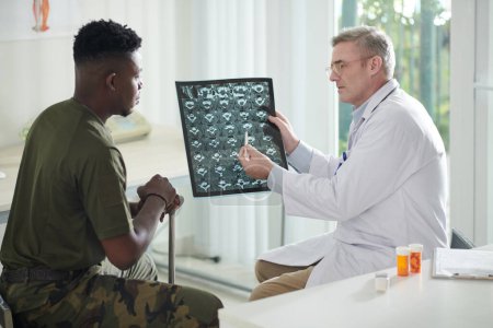 Photo for Serious doctor talking to military man about recovery progress after examining his spine MRI image - Royalty Free Image