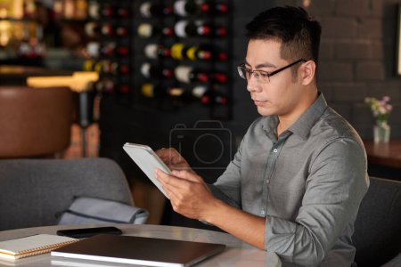 Téléchargez les photos : Homme d'affaires lisant un document sur une tablette numérique lorsqu'il travaille à table dans un café - en image libre de droit