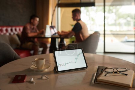 Photo for Stock market chart on digital tablet next to cup of coffee and planners on table, traders working in background - Royalty Free Image
