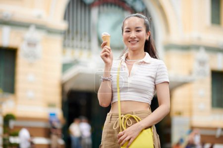 Téléchargez les photos : Portrait de jeune femme heureuse mangeant de la glace en marchant en ville - en image libre de droit