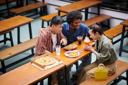 Foto de Diverso grupo de amigos bromeando y riéndose al tomar pizza y bebidas en la cafetería al aire libre - Imagen libre de derechos