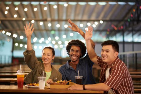 Foto de Sonriendo a los jóvenes sentados en la mesa de la cafetería y saludando amigo para atraer la atención - Imagen libre de derechos