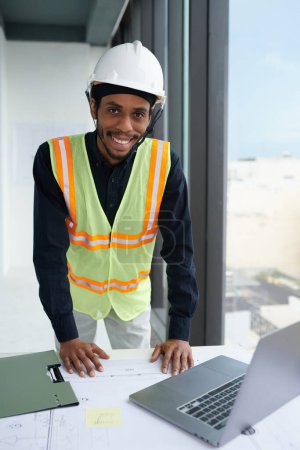Téléchargez les photos : Portrait de travailleur de la construction souriant en casque de sécurité appuyé sur le bureau avec plan de construction et ordinateur portable ouvert - en image libre de droit