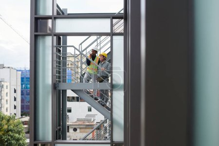 Téléchargez les photos : Ingénieur et contremaître debout sur les escaliers à l'extérieur du bâtiment et utilisant talkie-walkie pour communiquer avec l'équipe sur le chantier de construction - en image libre de droit
