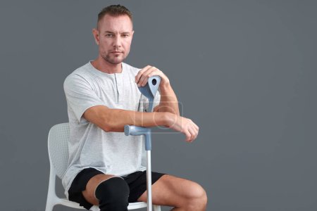 Photo for Mature man with hurt knees sitting on chair and leaning on crutch - Royalty Free Image