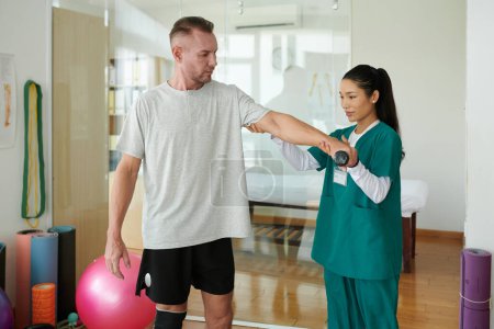 Foto de Trabajador del centro de rehabilitación que ayuda al cliente a hacer ejercicio con pesas para fortalecer los músculos del brazo - Imagen libre de derechos