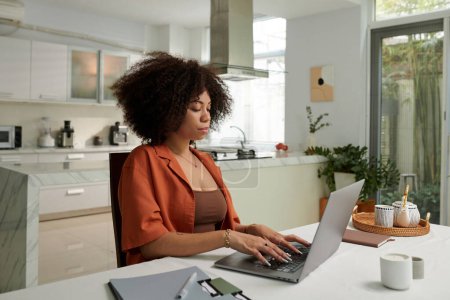 Téléchargez les photos : Femme sérieuse travaillant sur ordinateur portable à la table de cuisine à la maison - en image libre de droit