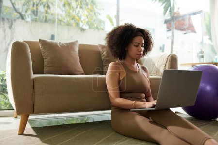 Foto de Mujer seria sentada en el suelo en casa y revisando correos electrónicos y mensajes de colegas en la computadora portátil - Imagen libre de derechos