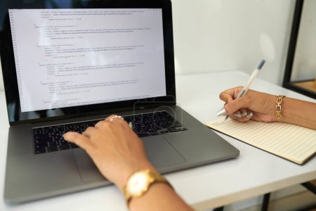 Téléchargez les photos : Femme examinant le code de programmation sur l'écran d'ordinateur portable et prenant des notes dans le planificateur - en image libre de droit