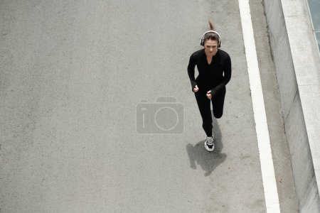 Foto de Vista de ángulo alto de la joven atlética en auriculares inalámbricos corriendo a lo largo de la calle a la música - Imagen libre de derechos