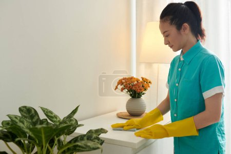 Téléchargez les photos : Nettoyant fille en uniforme et gants en caoutchouc essuyant la poussière de la table pendant son travail dans l'appartement - en image libre de droit