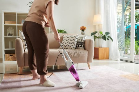 Foto de Primer plano de ama de casa utilizando aspiradora para aspirar la alfombra en la sala de estar durante las tareas domésticas en casa - Imagen libre de derechos