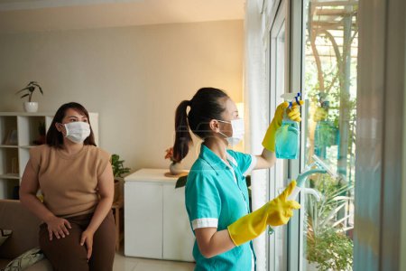 Téléchargez les photos : Jeune femme de service de nettoyage lave-vitres dans la chambre avec propriétaire regardant pour le processus - en image libre de droit