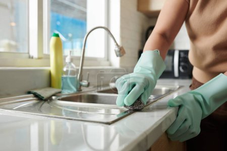 Close-up of housewife in protective gloves cleaning sink in kitchen during housework