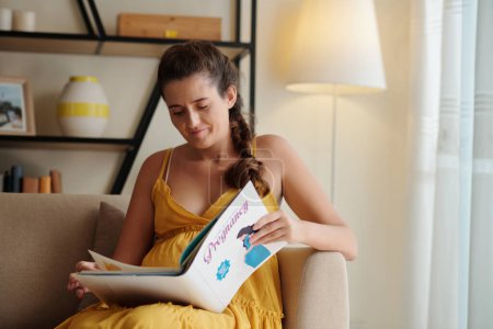 Foto de Mujer embarazada joven sentada en el sofá y leyendo libro sobre el embarazo durante su tiempo libre en casa - Imagen libre de derechos