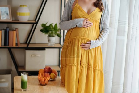 Téléchargez les photos : Gros plan de la femme enceinte en robe jaune caressant son ventre enceinte debout dans le salon - en image libre de droit