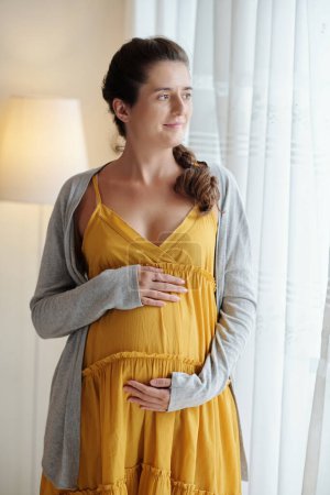 Foto de Mujer embarazada joven sosteniendo su vientre embarazada y mirando a la ventana con expresión pensativa mientras está de pie en la habitación - Imagen libre de derechos