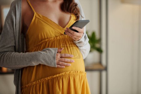 Téléchargez les photos : Gros plan de la femme enceinte caressant son ventre et lisant un message sur son téléphone portable - en image libre de droit