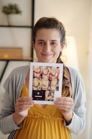 Foto de Retrato de mujer embarazada feliz mostrando la imagen de ultrasonido de su bebé y sonriendo a la cámara - Imagen libre de derechos