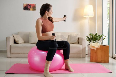 Foto de Mujer embarazada joven sentada en la pelota de fitness y haciendo ejercicio con pesas en casa - Imagen libre de derechos