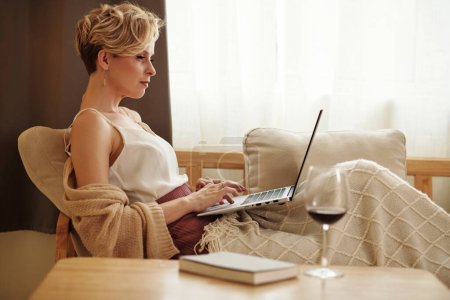 Photo for Side view shot of modern young adult Caucasian woman relaxing at home sitting on couch surfing Internet on laptop - Royalty Free Image