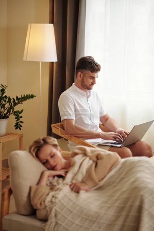 Photo for Vertical selective focus shot of young adult Caucasian man sitting in living room using laptop while his girlfriend taking nap - Royalty Free Image