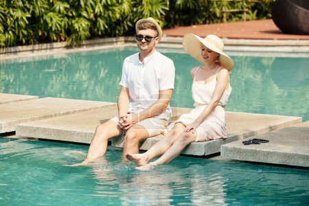 Photo for Modern young adult Caucasian man and woman sitting relaxed at pool with legs in water on hot summer day - Royalty Free Image