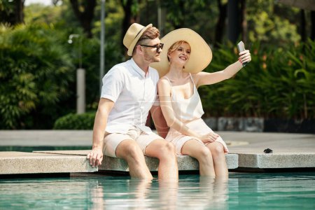 Photo for Young adult Caucasian couple enjoying spending time together at hotel territory sitting at pool with legs in water taking selfie on smartphone - Royalty Free Image
