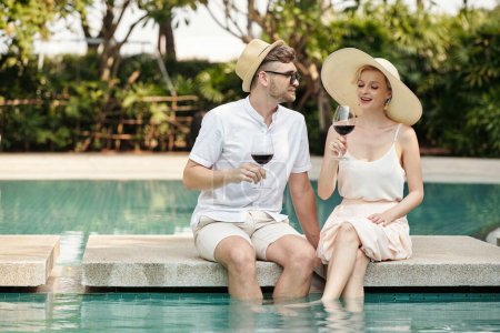 Téléchargez les photos : Élégant jeune homme et femme caucasien amoureux assis détendu à la piscine avec les jambes dans l'eau flirter et boire du vin rouge pendant les vacances - en image libre de droit