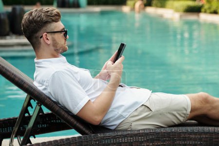 Photo for Side view of young adult Caucasian man relaxing on deck chair at poolside surfing Internet on smartphone on summer day - Royalty Free Image