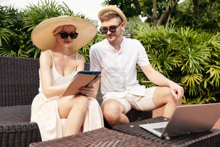 Téléchargez les photos : Jeune homme et femme caucasiens adultes assis à l'extérieur sur une journée ensoleillée d'été travaillant ensemble sur le projet en utilisant une tablette numérique et un ordinateur portable - en image libre de droit