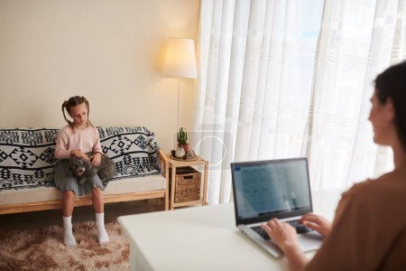 Téléchargez les photos : Mère travaillant sur un ordinateur portable à la maison lorsque sa fille joue avec le chien - en image libre de droit