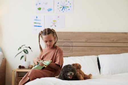 Photo for Preteen girl sitting on bed and playing ukulele for her little dog - Royalty Free Image