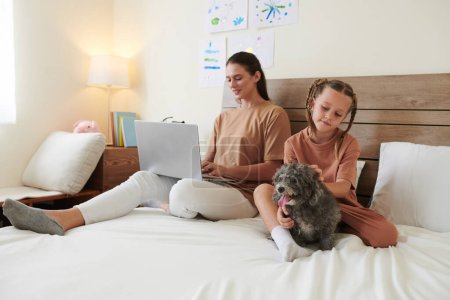 Téléchargez les photos : Jeune femme travaillant sur un ordinateur portable au lit lorsque la fille joue petit ours chien par - en image libre de droit