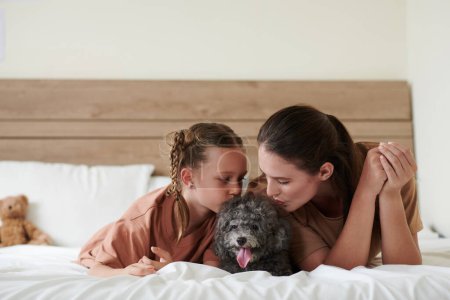 Téléchargez les photos : Mère et fille embrassant petit chien sur la tête en se reposant sur le lit à la maison - en image libre de droit