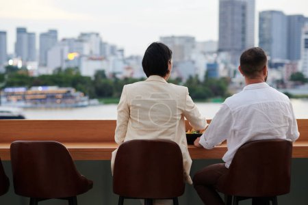 Téléchargez les photos : Couple d'affaires ayant des bières froides et des collations et regardant beau paysage urbain devant bar sur le toit après le travail - en image libre de droit