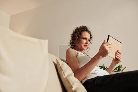 Foto de Elegante mujer de mediana edad sentada en el sofá y viendo un nuevo programa popular en la tableta digital - Imagen libre de derechos